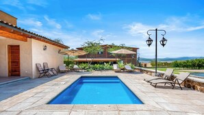 Una piscina con sillas de descanso y sombrillas, rodeada por un patio de piedra y una casa con techo de tejas.