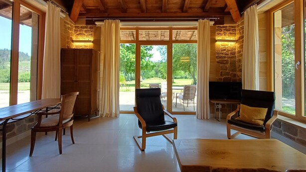 A cozy room with a wooden ceiling, stone walls, and large windows. There are two wooden chairs, a table, and a TV.