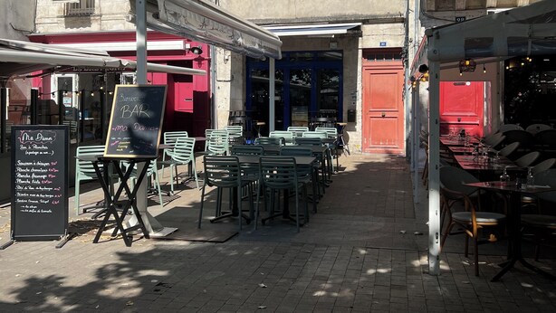 Un café al aire libre con mesas y sillas, un menú en la pizarra y un edificio con varias ventanas y contraventanas al fondo.