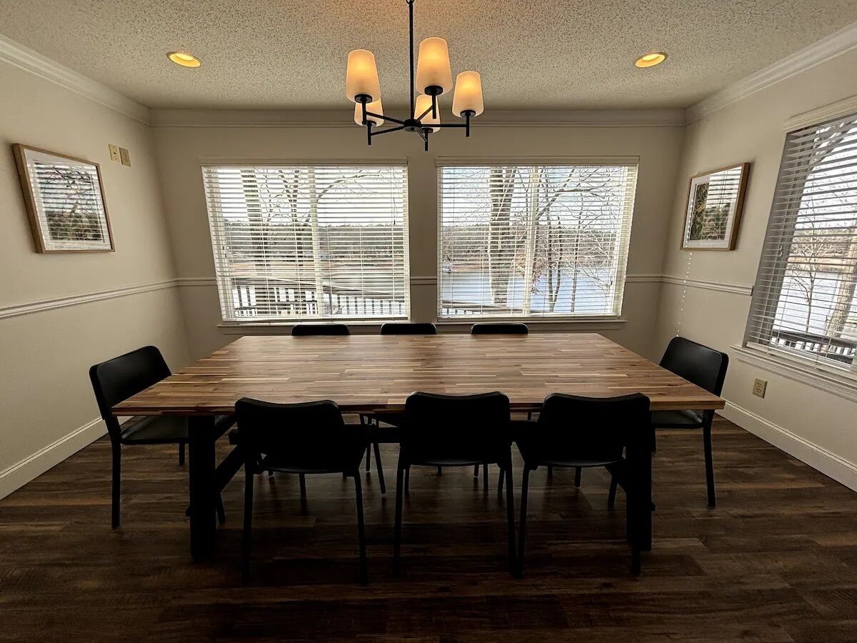 A conference room with a large wooden table, six chairs, a chandelier, and large windows with blinds.