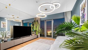 A modern living room with a large flat-screen TV, a sofa, and a coffee table. There are plants and a city view through the windows. - Dubai Marina 3BR in Vida | Pool, Gym, Balcony, Walk Everywhere (Dubai)
