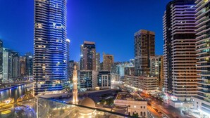 Une vue nocturne d’une ville, avec des immeubles modernes de grande hauteur, une mosquée imposante surmontée d’un dôme, et une rue animée où la circulation est dense.