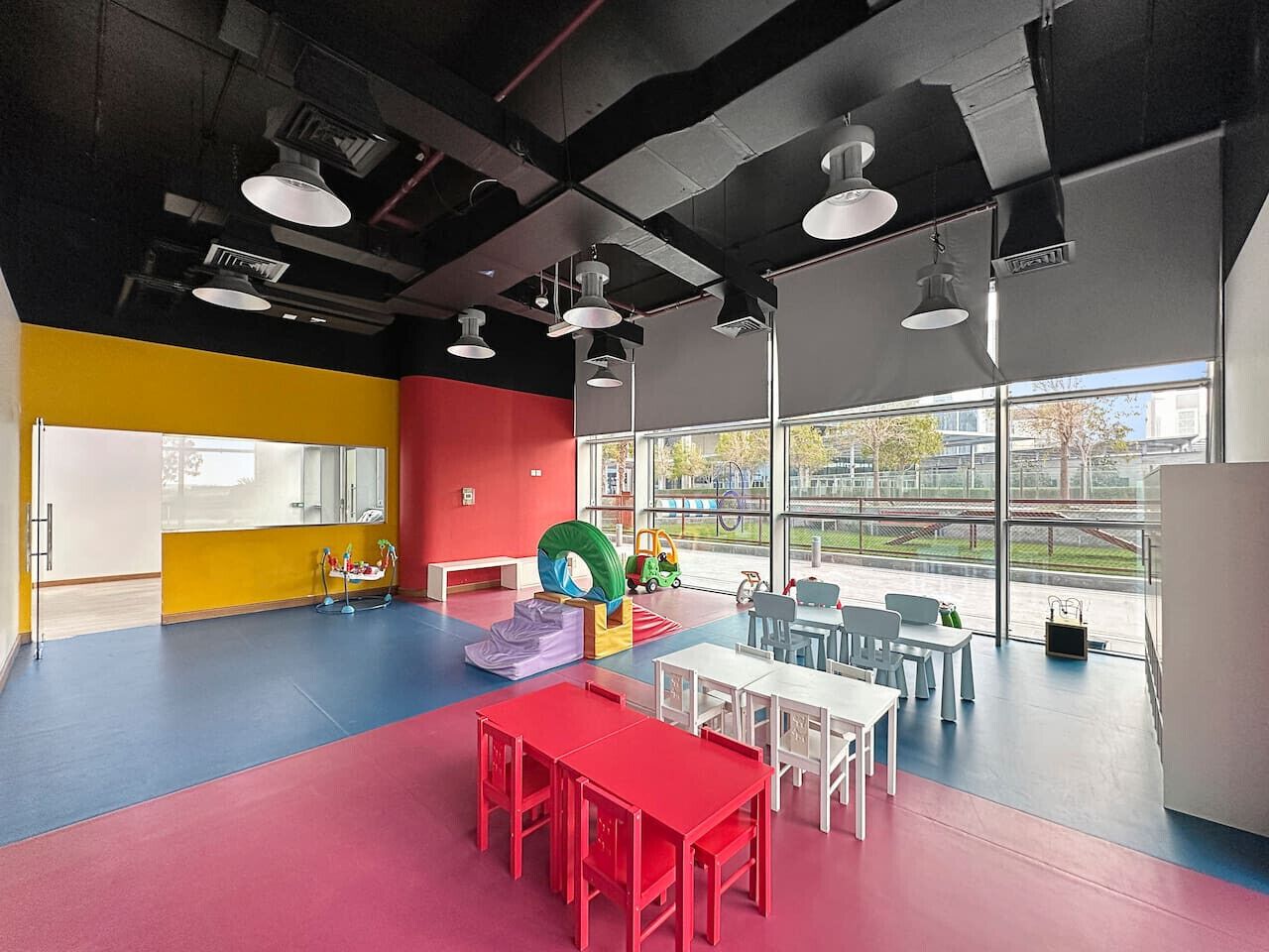 Un espacio interior, lleno de color, con área de juegos, mesas y sillas, ventanas amplias y vistas al exterior.