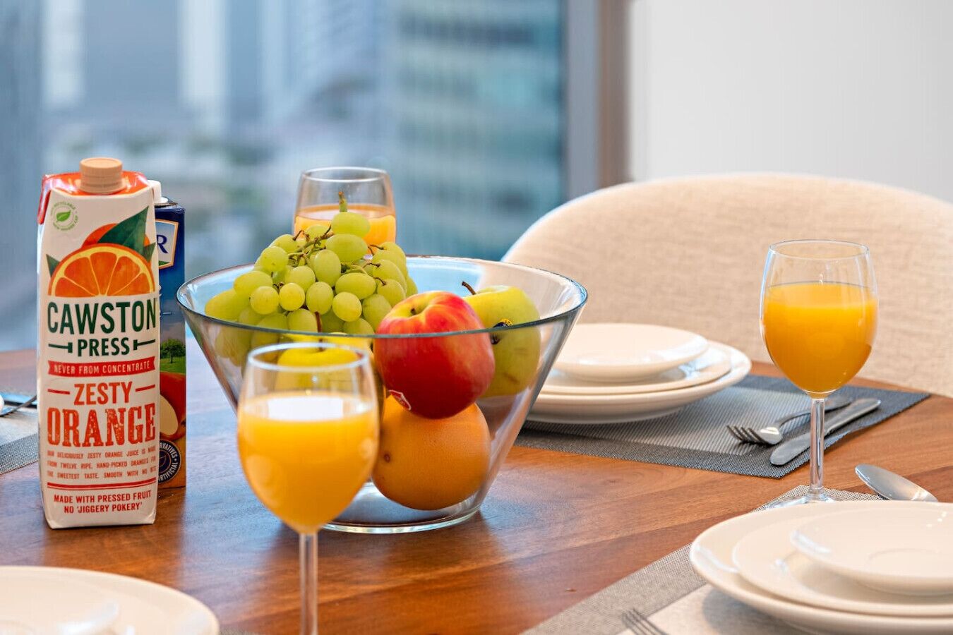 Une table mise pour le petit-déjeuner, avec un panier de fruits contenant des raisins, des pommes et des oranges, un verre de jus d’orange et un emballage de jus d’orange pétillant.