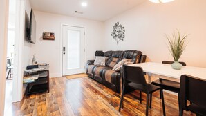 A living room with a sofa, a dining table with chairs, a TV mounted on the wall, and a door.