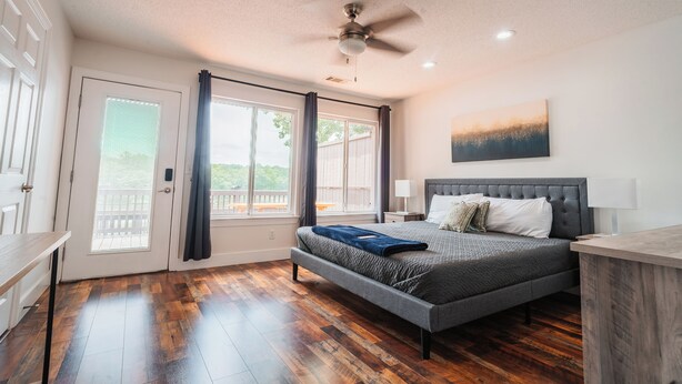 A bedroom with a bed, a ceiling fan, a window with curtains, a door, and a view of the outdoors.