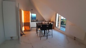 Een ruime zolderkamer met een eettafel en stoelen, grote ramen en een keukenhoek.