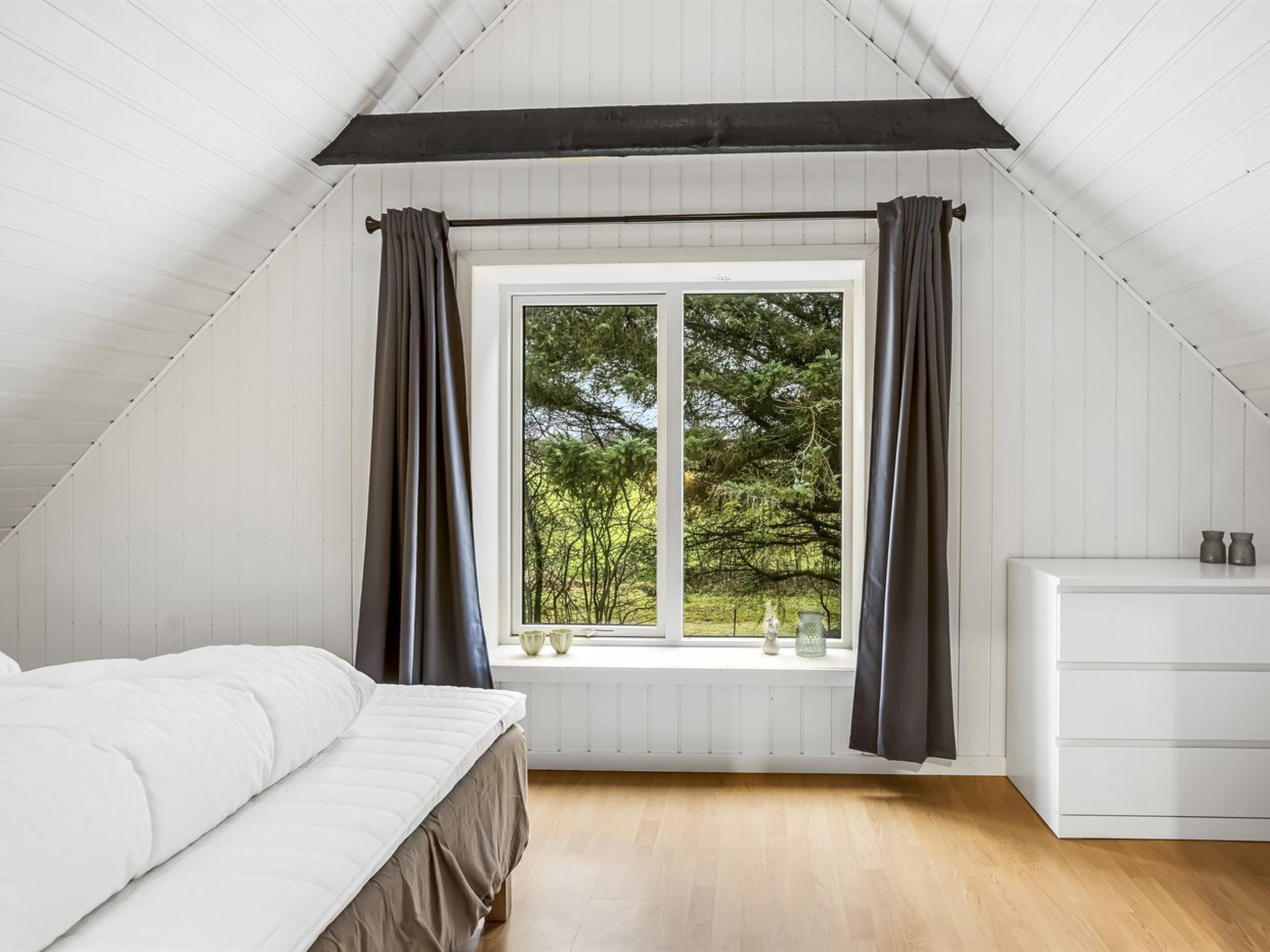 A bedroom with a sloped ceiling, a window with curtains, a bed, and a dresser.