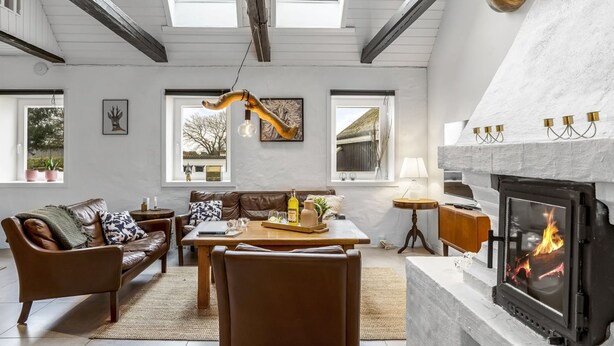 A cozy living room with a fireplace, a sofa, a dining table, and a view of the outdoors through large windows. - "Thorgot" - 3km from the sea by Interhome (Pandrup)