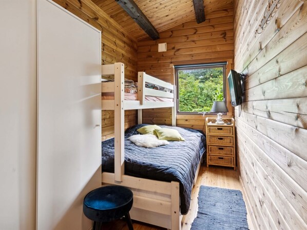 A compact wooden cabin room with bunk beds, a small bedside table, a lamp, and a window with a view of greenery.