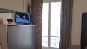 A modern living room with a kitchen area, a dining table, and a sofa. There is a television on a cabinet, and a window with curtains.