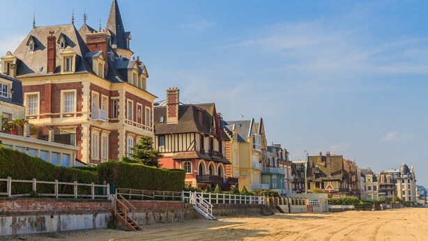 A coastal street with historic buildings, a sandy path, and a white fence.