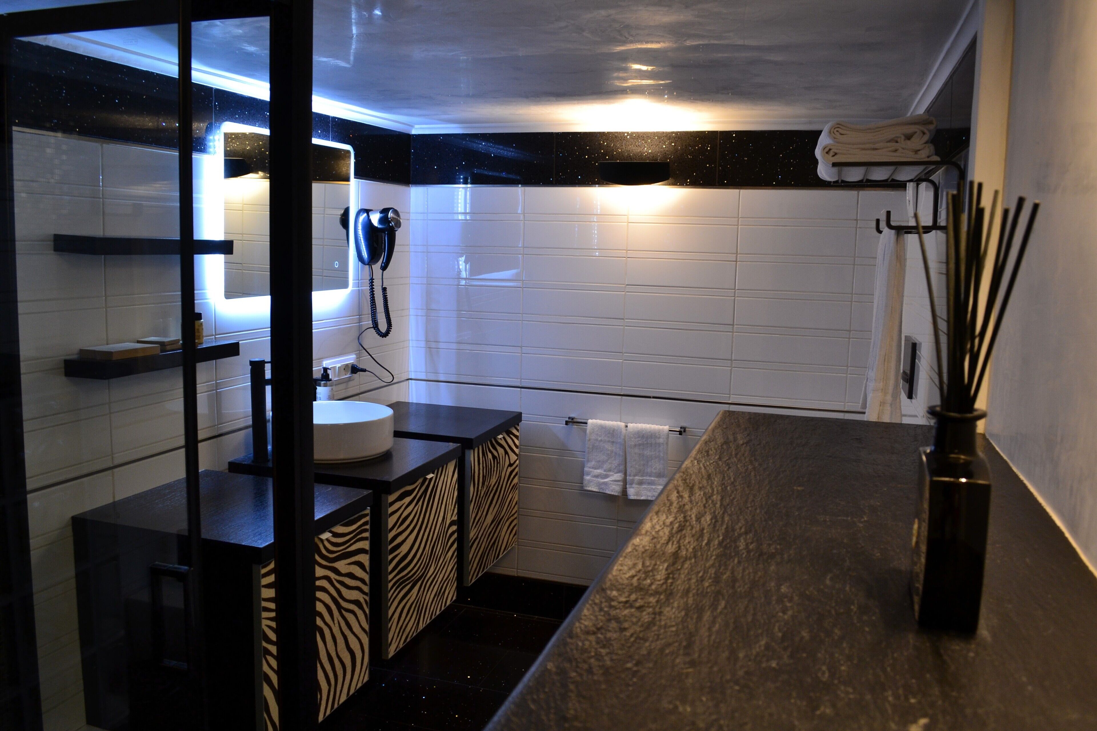 Un baño moderno con encimera negra, azulejos blancos, un mueble con estampado de cebra, un secador de pelo y un perchero.