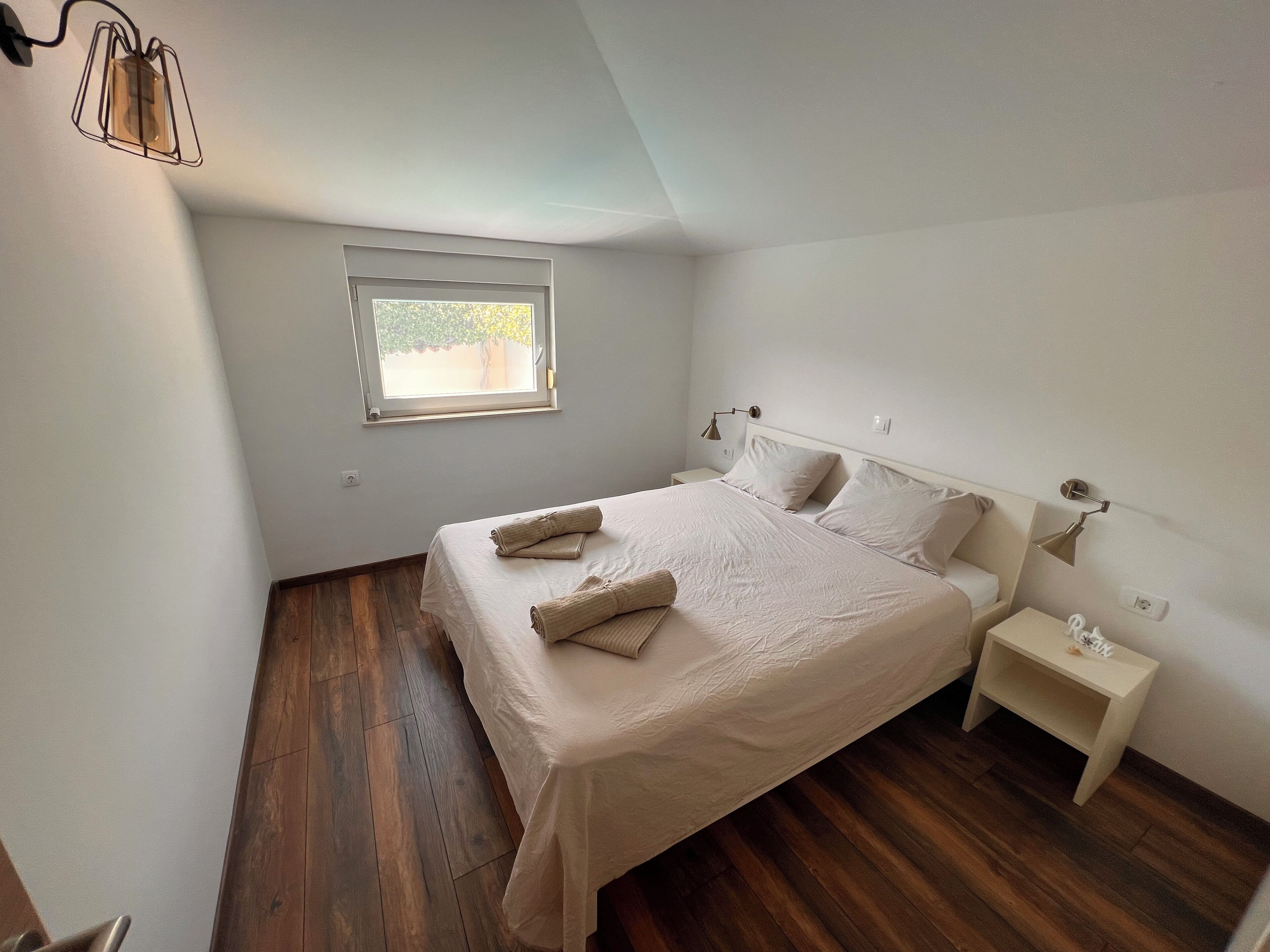 Un dormitorio con cama, mesita de noche, lámpara, ventana y suelo de madera.