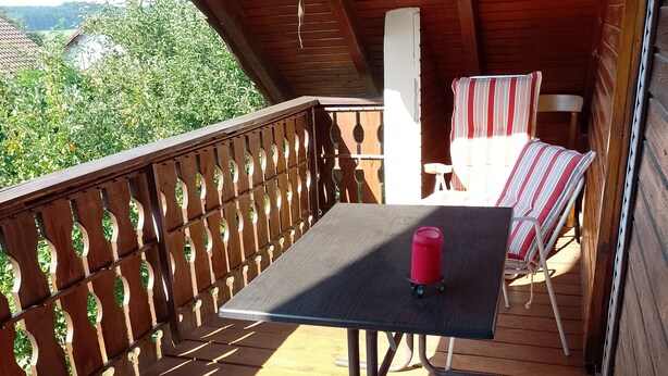 Une terrasse en bois avec une table et deux chaises à rayures, un carillon éolien suspendu et, en arrière-plan, une vue sur la verdure.