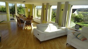 A spacious living room with a wooden floor, a white sofa, a dining table with chairs, and large windows offering a view of greenery and hills.