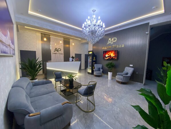 A modern lobby with a reception desk, seating area, chandelier, and a TV displaying a fire.