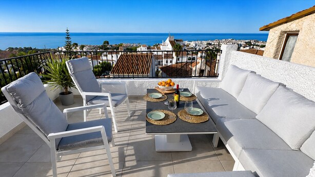 Une terrasse avec une table dressée pour deux personnes, des meubles d’extérieur blancs, une plante en pot et une vue sur la mer et la ville.