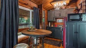 A cozy cabin interior with a wooden dining table, bar stools, a kitchen area with a microwave and cabinets, and a large window with curtains.