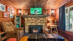 A cozy cabin interior with a stone fireplace, a television mounted above it, and a comfortable seating area with a leather sofa and ottoman.