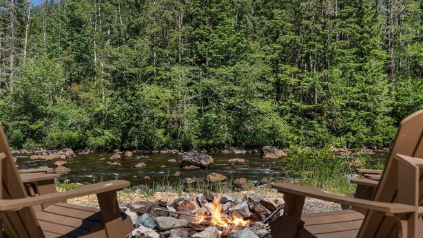A campfire with rocks, surrounded by wooden chairs, set against a backdrop of dense forest and a flowing river.
