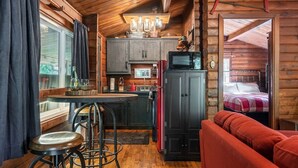 A cozy cabin interior with a kitchen area, a dining table with stools, a red sofa, and a bed with a checkered blanket.