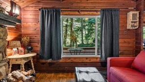 A cozy cabin interior with a wooden log wall, a window with curtains, a red sofa, a small table with a lamp, and a view of the forest outside.