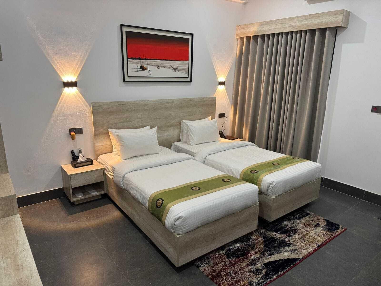 A double bed with two pillows, a bedside table with a phone, a framed picture on the wall, and a window with curtains.
