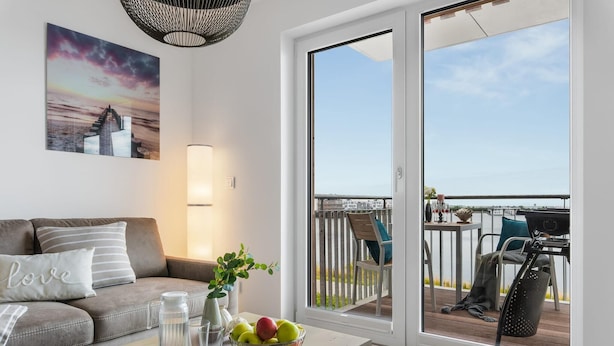 Un salon moderne avec un canapé, une table sur laquelle se trouve un panier de fruits, un vase et une lampe. On aperçoit un balcon avec une rambarde et des meubles d’extérieur à travers une porte-fenêtre coulissante.