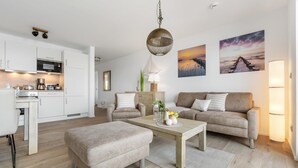 Un salon moderne avec un canapé, un pouf et une table basse, une cuisine avec des meubles blancs et des appareils électroménagers, et deux tableaux représentant un coucher de soleil accrochés au mur.