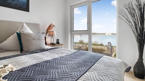 Une chambre moderne avec un grand lit, une table de chevet, une lampe et une vue sur une étendue d’eau, visible à travers une grande fenêtre.