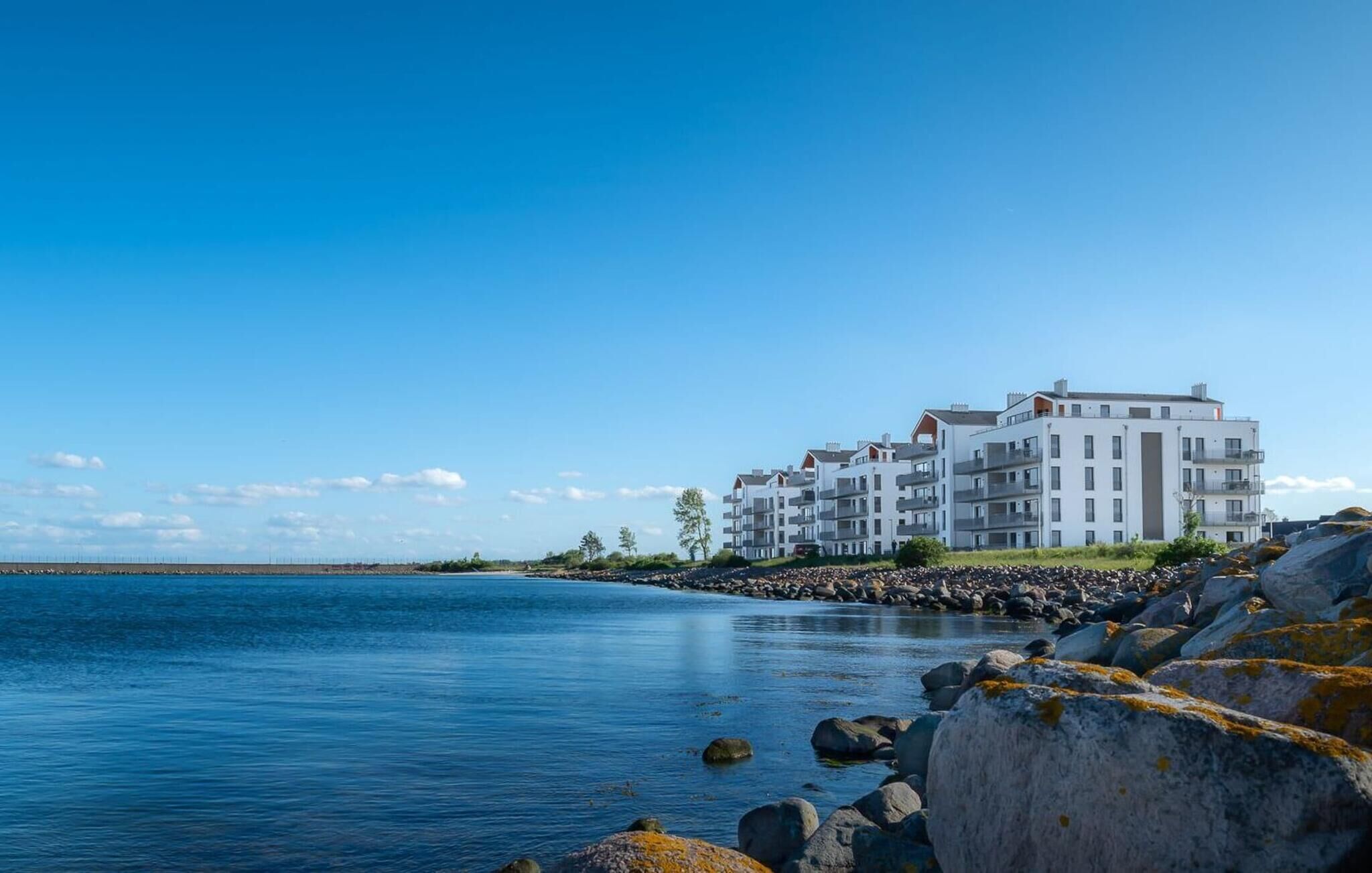 A coastal building with multiple floors, likely a hotel, situated near a calm body of water with visible rocks and sparse vegetation.