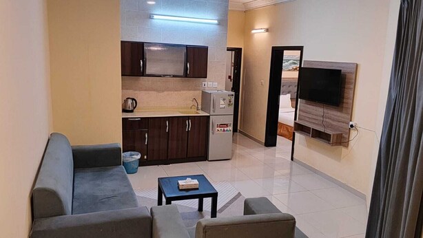 A compact living space with a kitchenette, a sofa, a small table, and a TV mounted on the wall. - Wadan Al-Mutamayeza Furnished Apartments (Rabigh)