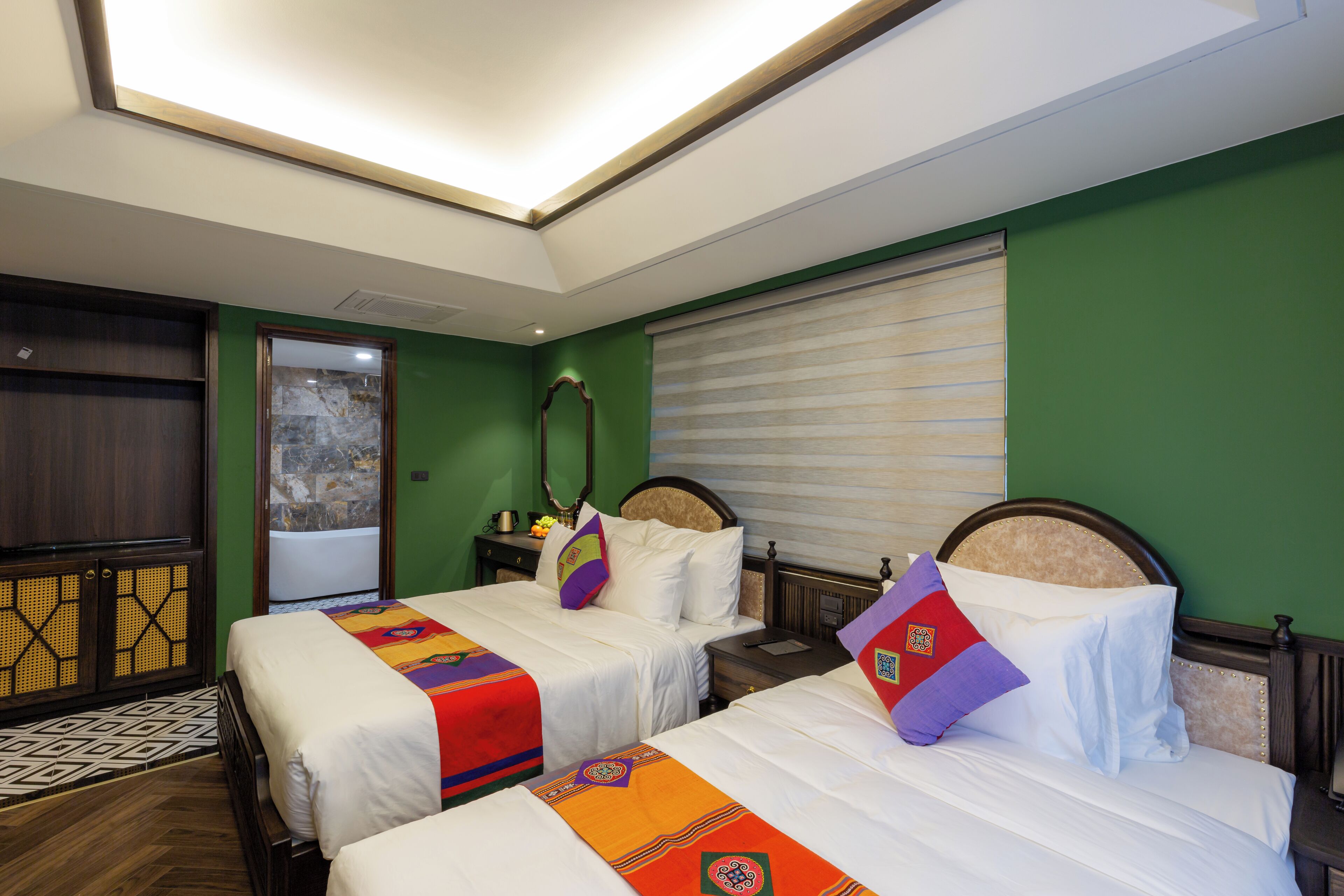 A hotel room with two beds, a large window with blinds, a wooden cabinet, and a patterned rug.