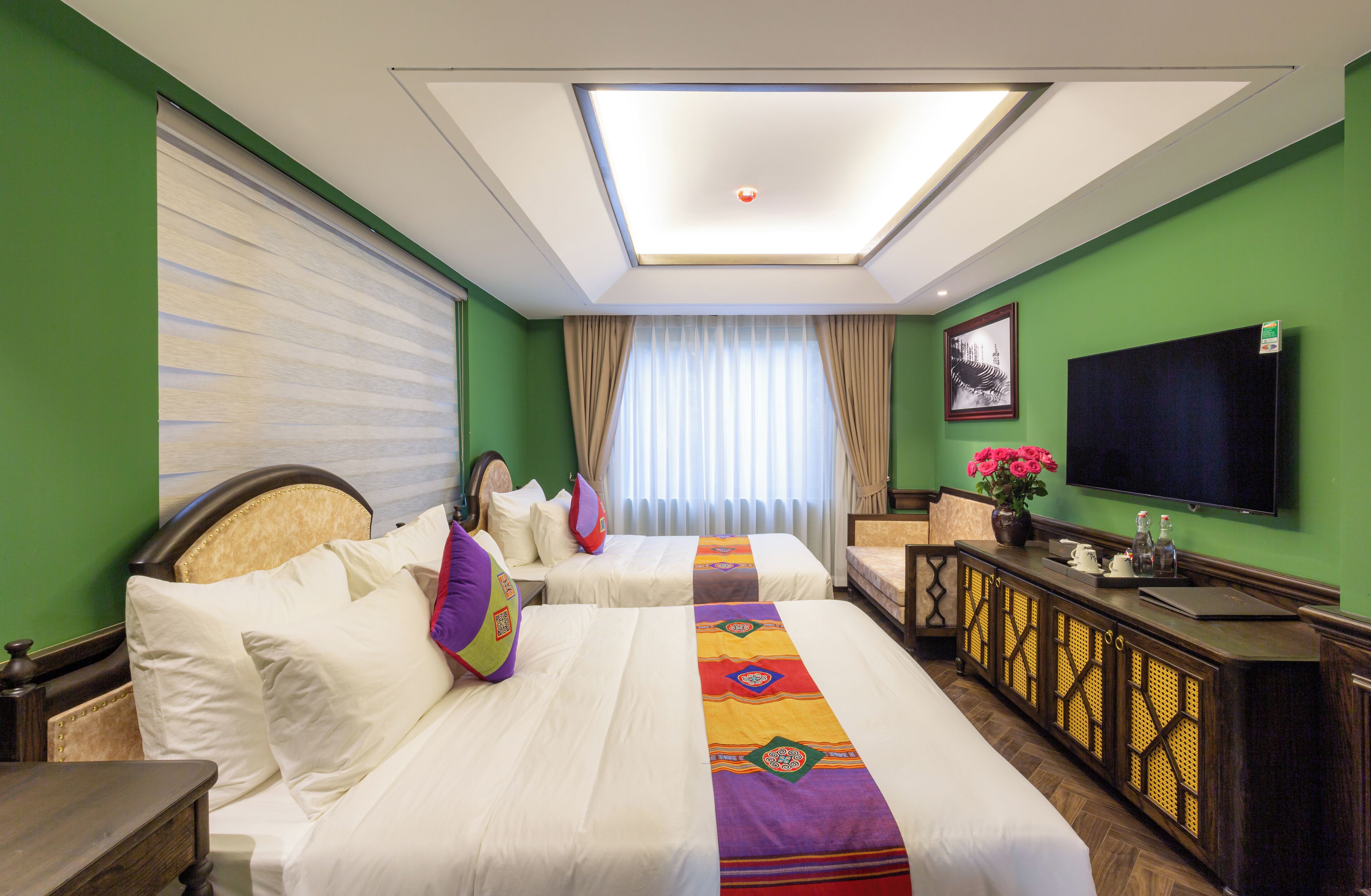 A hotel room with two beds, a television, a desk with a vase of flowers, and a window with curtains.
