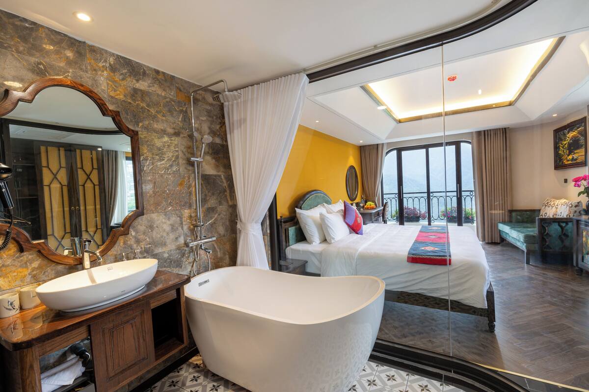 A modern hotel room with a large bathtub, a separate shower area, a large bed, and a balcony with a view.