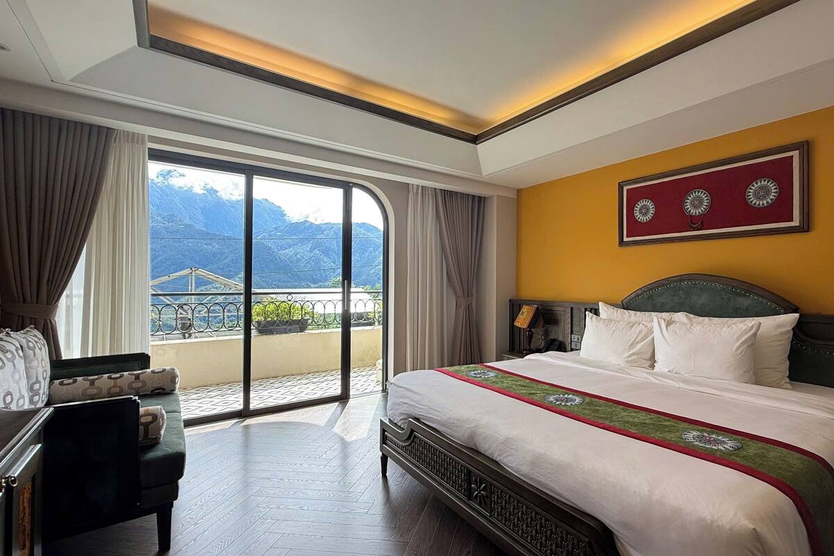 A hotel room with a large bed, a desk, and a view of mountains through the balcony.