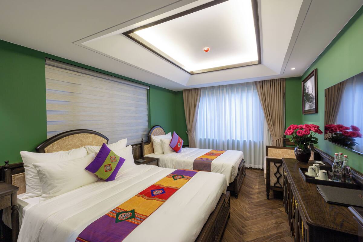 A hotel room with two beds, a desk, a chair, a small table, a vase with flowers, and a window with curtains.