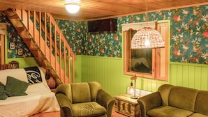Una habitación acogedora con una escalera de madera, paredes verdes, papel tapiz floral, una cama con almohadas verdes, dos sillones verdes, una mesita de madera con una lámpara y una ventana con marco de madera.