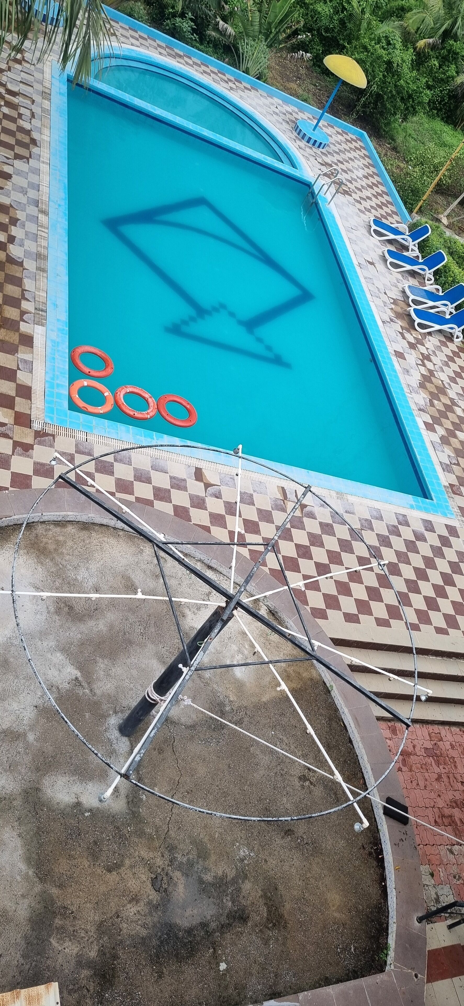 A swimming pool with a checkered pattern around the edge, a bicycle leaning against the poolside, and a pair of lounge chairs with an umbrella.