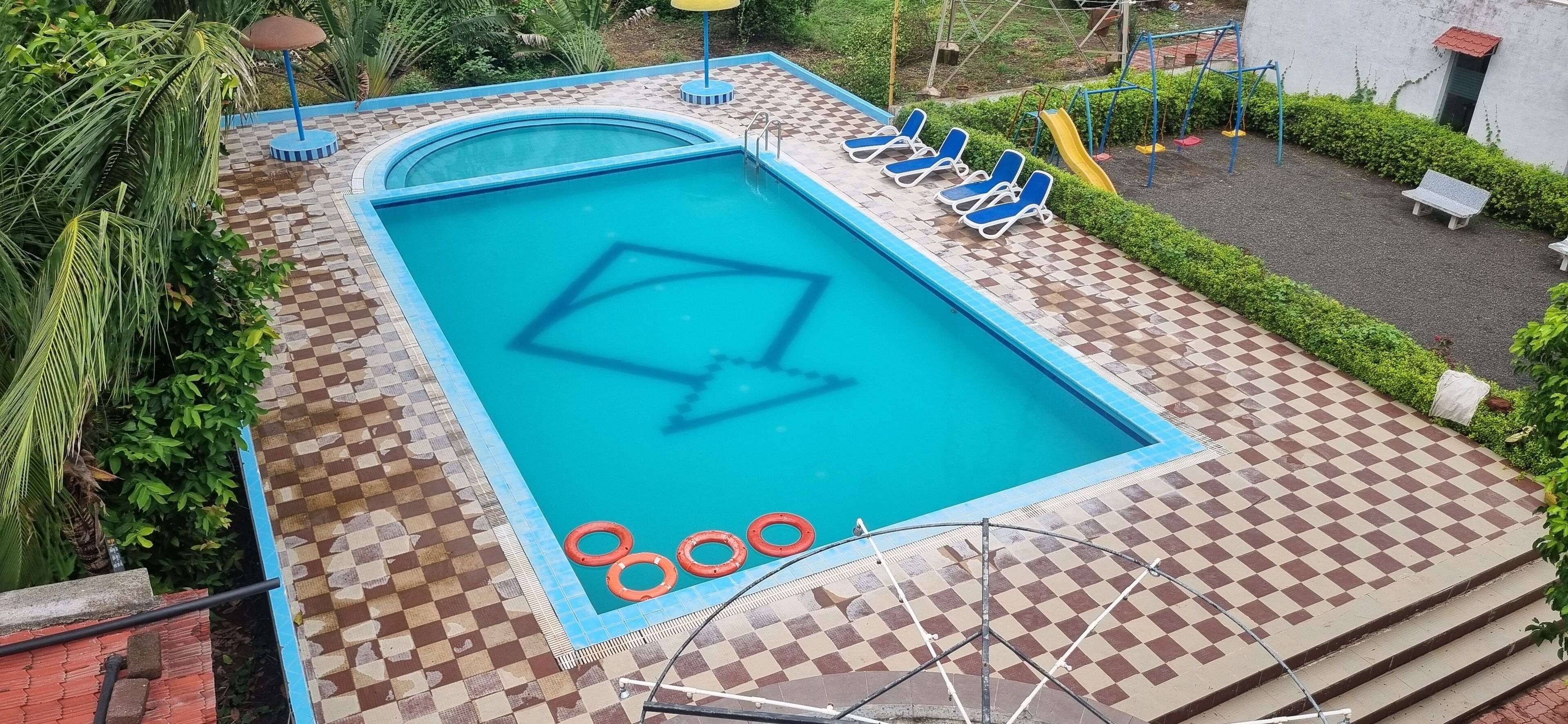 Una piscina rectangular con un diseño de tablero de ajedrez a su alrededor, bordeada por sillas de descanso y un tobogán.