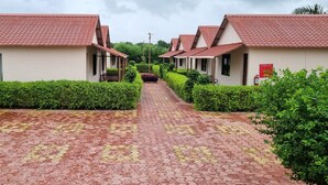 A pathway leading to a row of small, single-story buildings with red-tiled roofs, surrounded by well-maintained greenery. - PATANG FAMILY RESORT (Talala)