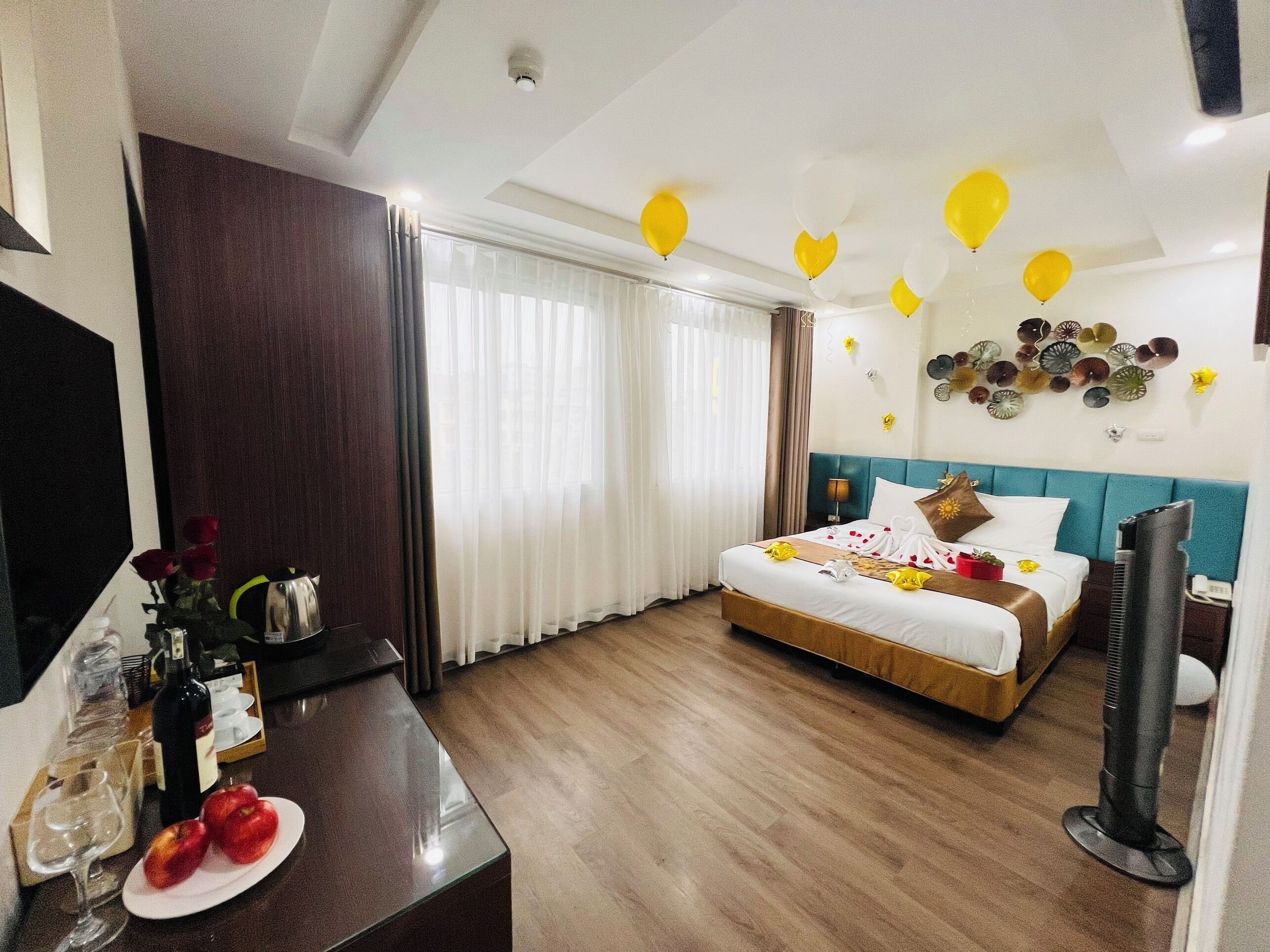 A hotel room with a bed, a TV, a desk with a coffee maker, and a dining table with apples and wine glasses.