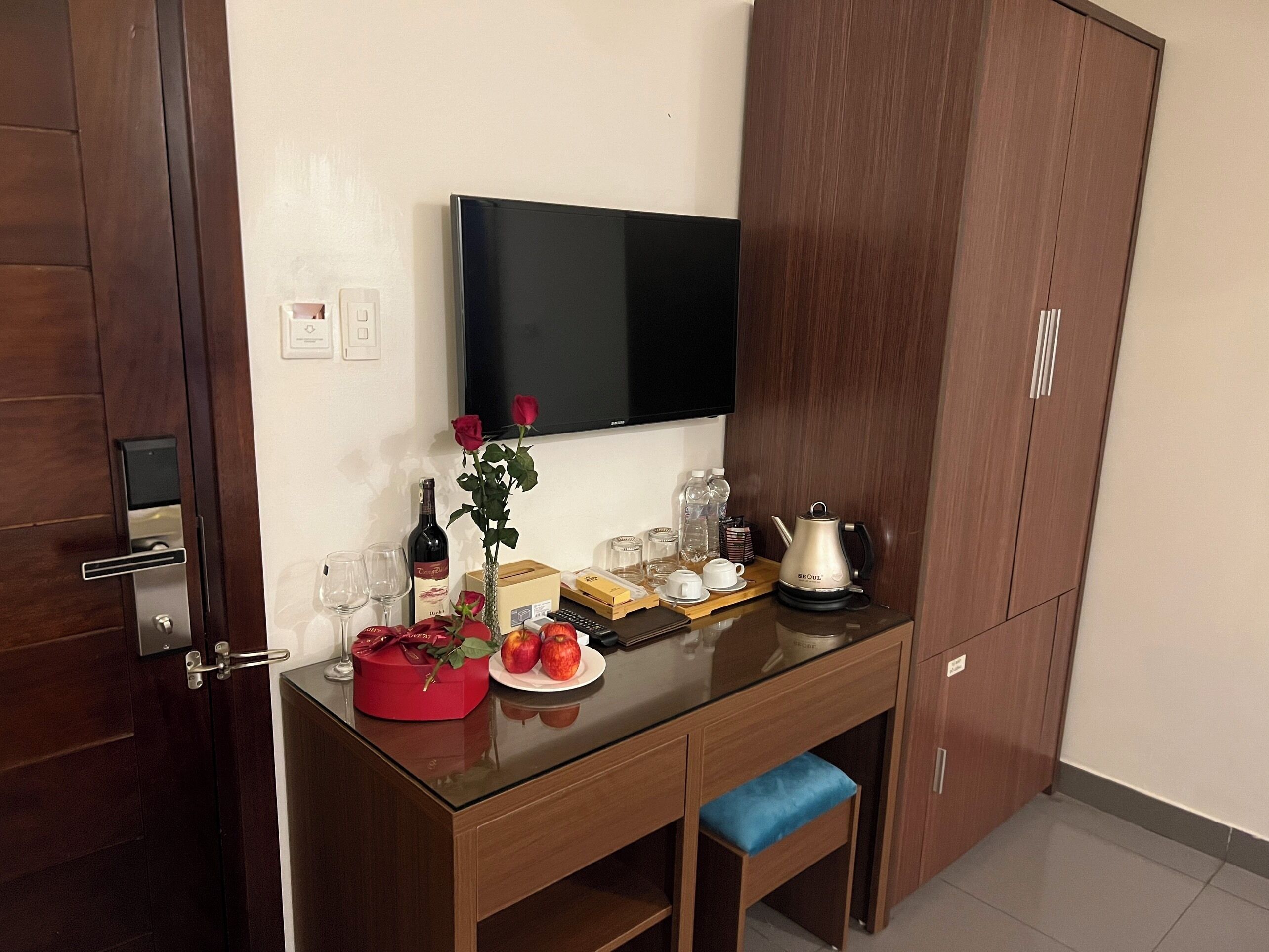 A compact hotel room with a wooden desk, a flat-screen TV, a minibar, and a small seating area.