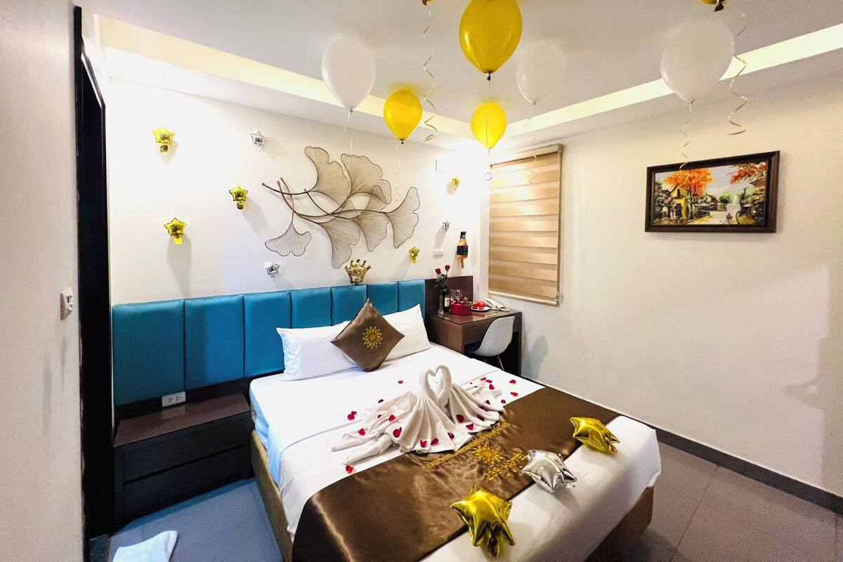 A hotel room with a bed, a bedside table, a window with blinds, and a wall decoration.