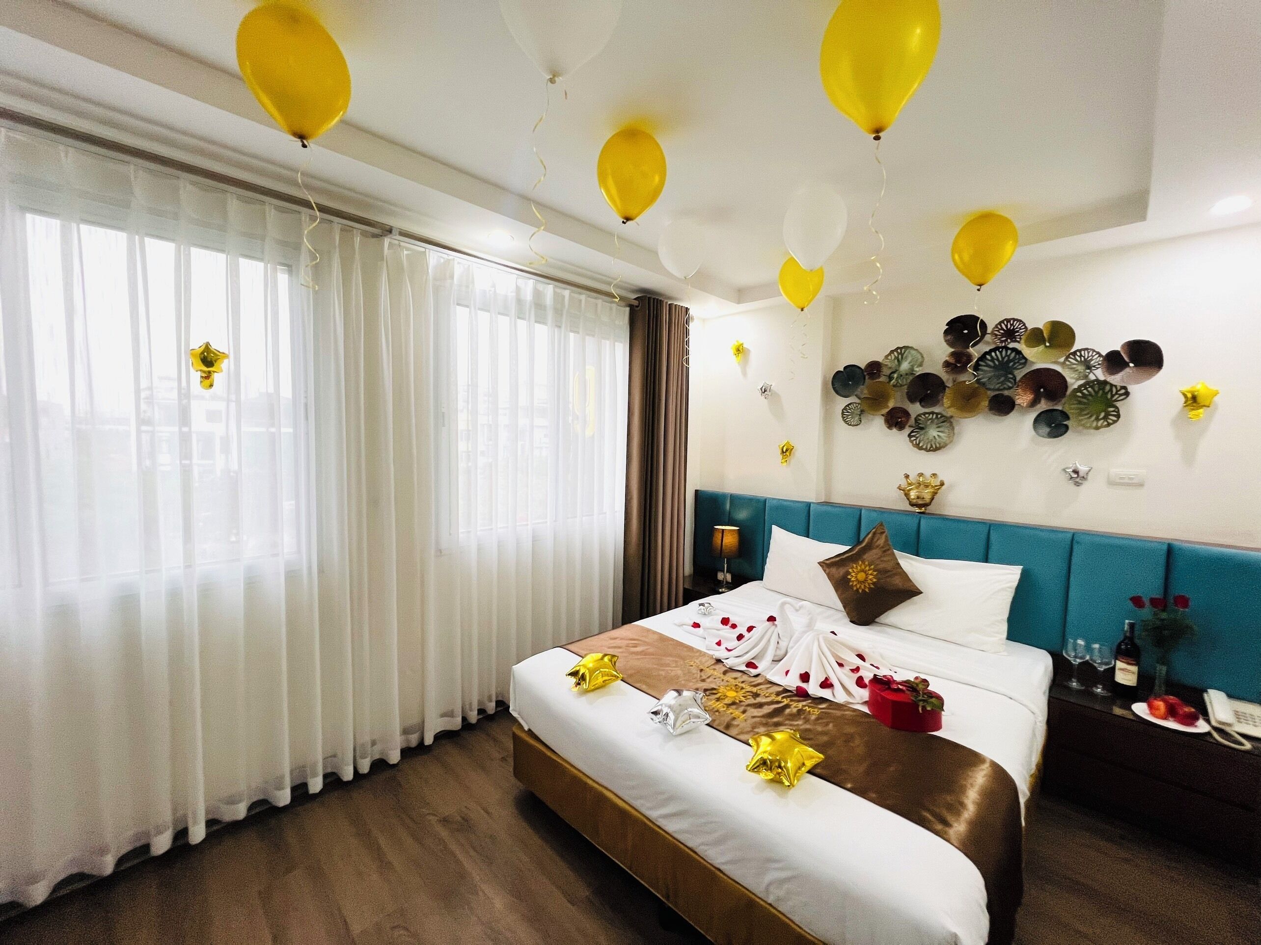 Una habitación de hotel con una cama grande, cortinas, un cabecero azul, decoración en la pared y globos.