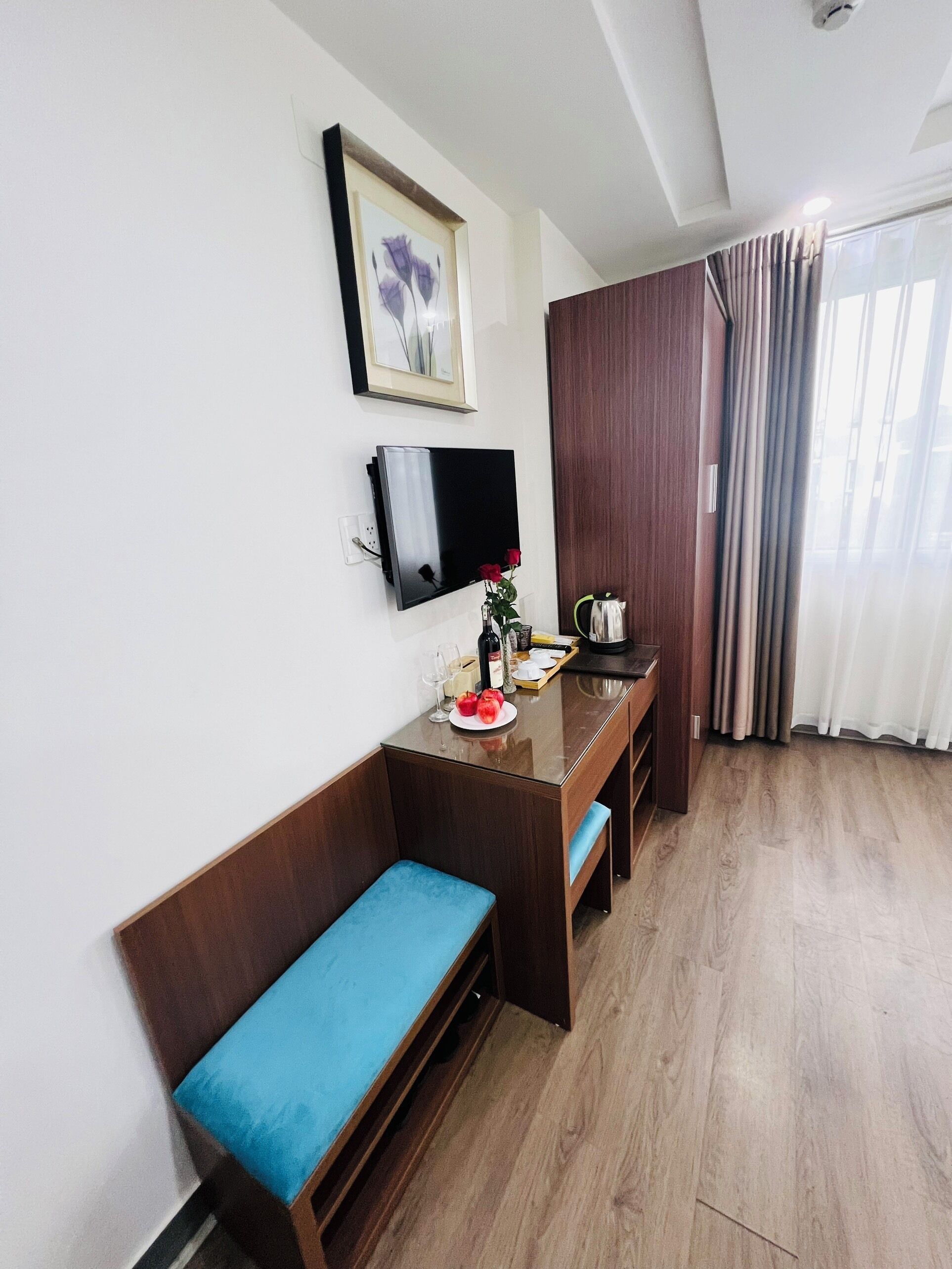 A compact room with a wooden bench, a small table with a vase of flowers, a flat-screen TV mounted on the wall, and a window with curtains.