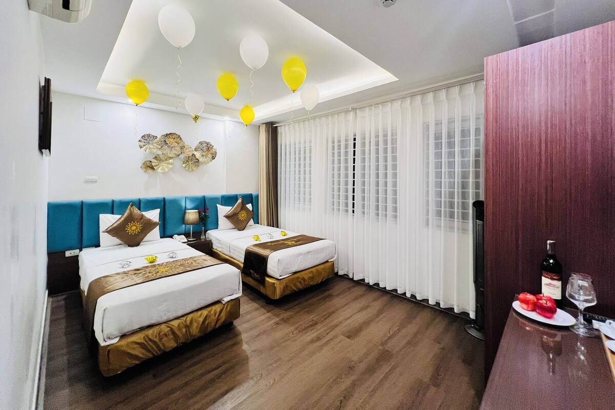 A hotel room with two beds, a wooden floor, a ceiling light, and a window with curtains.