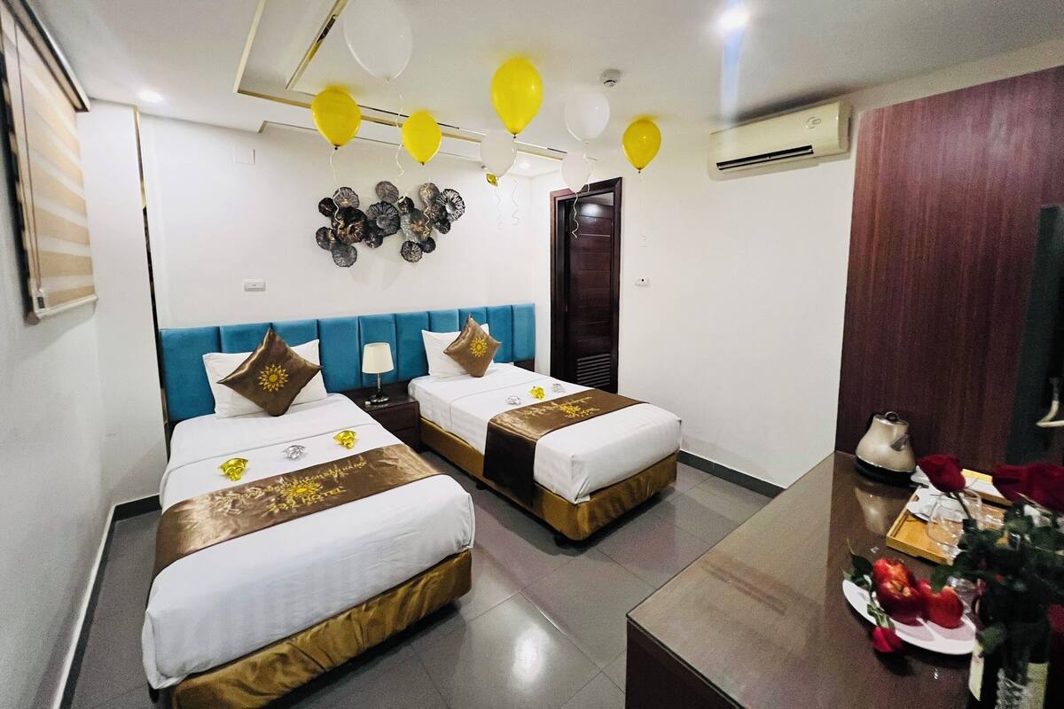 A hotel room with two beds, a wooden headboard, a wall decoration, and a dining table with a red rose.