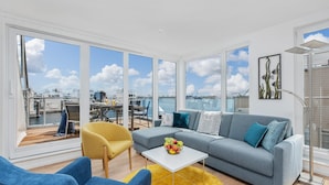 Un salon moderne avec un canapé gris, des fauteuils bleus et un fauteuil jaune. Une table blanche sur laquelle est posé un saladier de fruits se trouve au centre de la pi èce. De grandes fenêtres offrent une vue sur le port de plaisance et les bateaux.